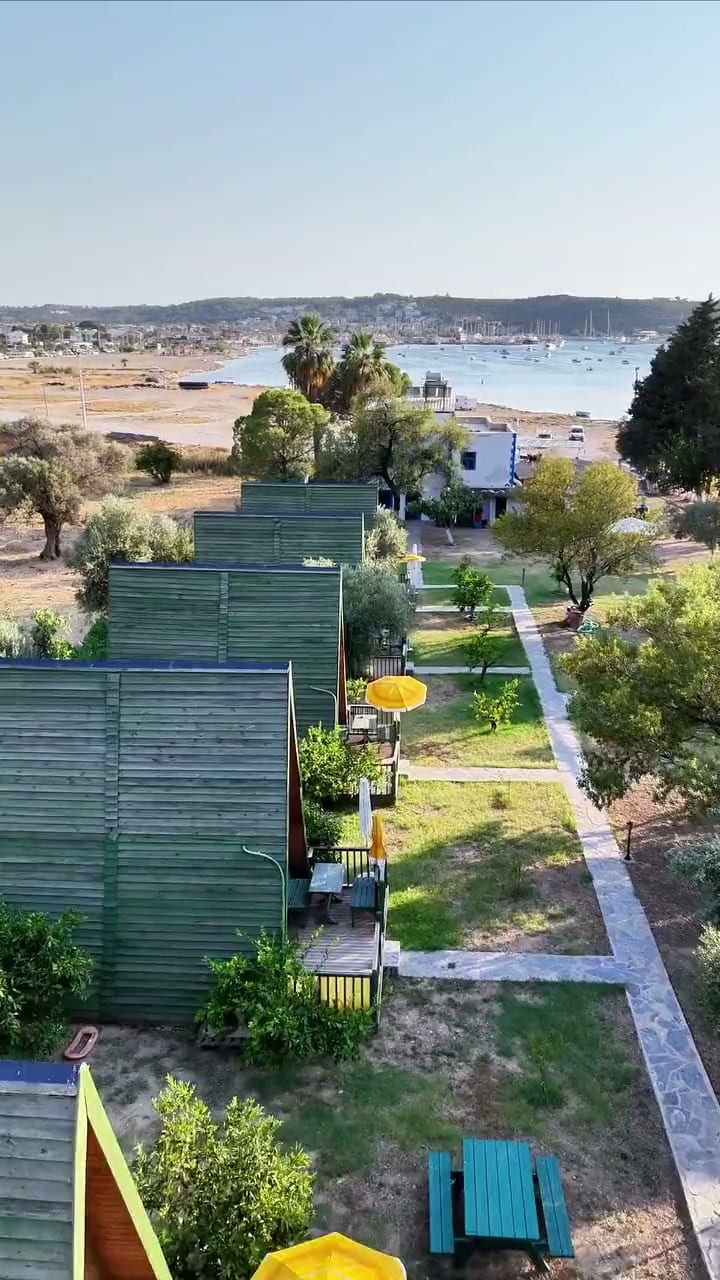 Sakin’in bahçesi, havadan: yeşil ahşap yapılar, taş yollar, zeytin ve mandalina ağaçları; arkada teknelerin durduğu körfez.