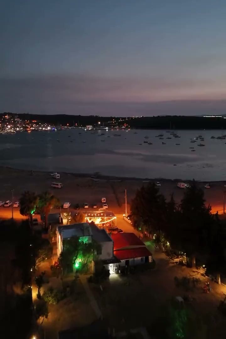 Akşam, körfez havadan: karşı yakada yerleşim ve suda tekneler; önde ışıkları yanan Sakin.
