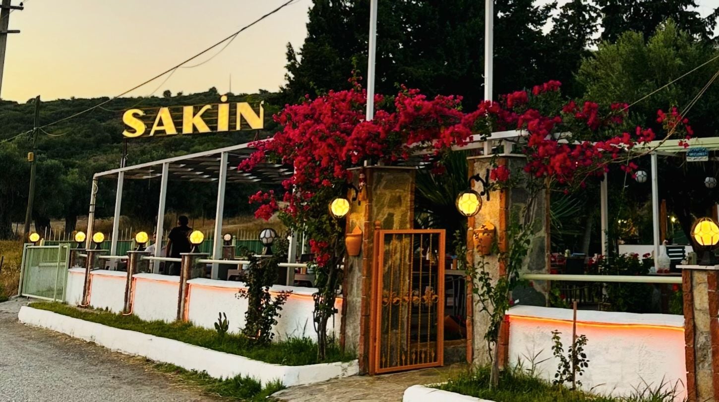 Gün batımında Sakin’in girişi: ışıklı tabela, begonviller ve fenerler.